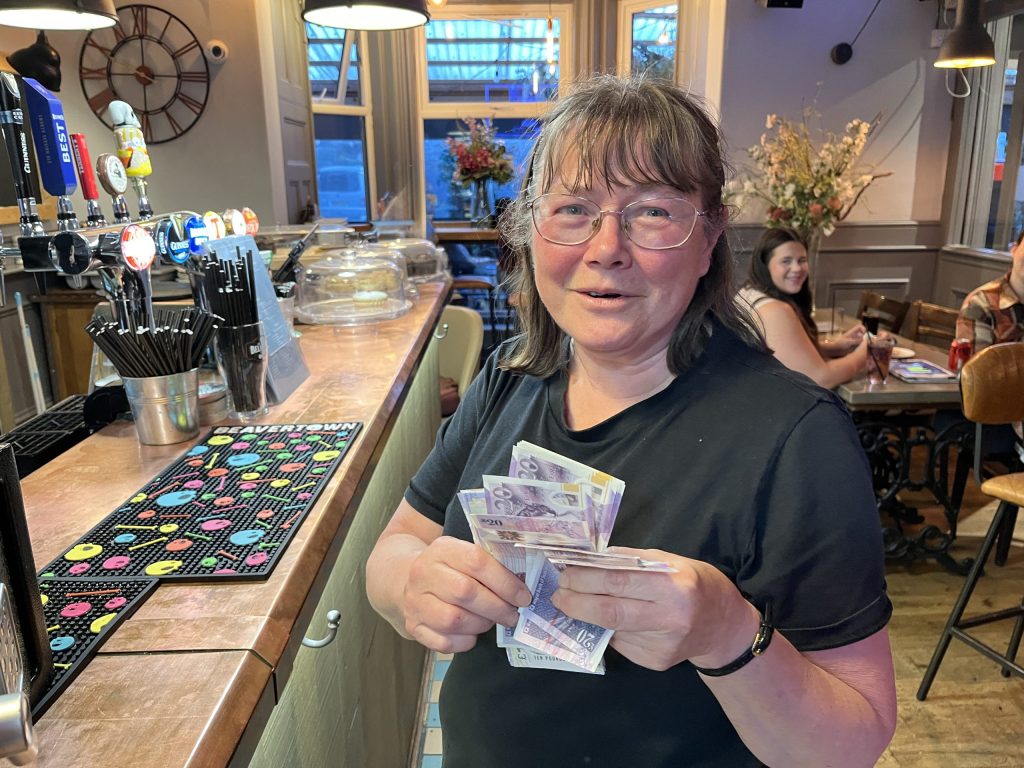 Smiling woman at a bar counter counting cash, with beer taps and a menu nearby in a cozy cafe setting.